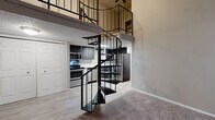 This loft-style space is defined by its dramatic vertical design, anchored by a sleek black metal spiral staircase that connects the main level to an open upper loft. The soaring ceilings in the main living area create an airy, open atmosphere, with the loft level overlooking the space below via a black metal railing. The vaulted ceiling in the loft features a textured finish, adding architectural character to the upper level.

The kitchen is equipped with stainless steel appliances, including a range, microwave, and refrigerator, complemented by contemporary dark cabinetry and light countertops. Light wood-style flooring runs throughout the kitchen and connects seamlessly to the main living area, which transitions to plush carpeting.

The bedrooms are carpeted and offer generous storage through double sliding closet doors. Natural light enters through windows fitted with blinds. Two bathrooms serve the space: one features a tub-shower combination with white tile surround, while the second includes a walk-in glass shower. Both bathrooms are finished with marble-style countertops, dark wood-tone vanity cabinetry, and wood-look flooring, providing a cohesive and modern aesthetic throughout.