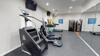 This single-floor fitness center offers a practical and functional workout environment designed to meet a variety of exercise needs. The space is outfitted with a lineup of cardio equipment, including a stair climber and multiple treadmills, positioned along the main floor area to maximize usability. Rubber flooring throughout provides both safety and durability underfoot.

Two wall-mounted flat-screen TVs are strategically placed to keep users entertained during workouts—one positioned high on the front wall and another mounted at eye level along the side wall. Recessed ceiling lighting ensures the room is well-lit, while natural light filters in through a window along the side wall, contributing to a bright and open atmosphere.

Exercise accessories, including medicine balls and a yoga mat, are neatly stored near the equipment, adding versatility for stretching and strength training routines. A posted fitness center rules board keeps guidelines visible to all users. Rounding out the amenities, a snack and beverage vending machine is conveniently located near the entrance, offering a quick refreshment option post-workout.