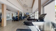 This single-level fitness center is thoughtfully designed to support a comprehensive workout experience within a bright, open environment. The space is defined by its dramatically high ceilings, which are accented by a stylish geometric chandelier and ceiling fans that enhance air circulation throughout. Large floor-to-ceiling windows and glass doors line one wall, flooding the interior with abundant natural light and offering views of a lush, palm-lined outdoor area, creating an airy and motivating atmosphere.

The flooring throughout is a durable wood-style material that lends warmth and visual continuity to the space. Full-length mirrors along the walls expand the sense of space and allow users to monitor their form during workouts. Wall-mounted flat-screen televisions are positioned for easy viewing from cardio equipment.

The cardio zone is well-stocked with multiple treadmills, elliptical trainers, and other aerobic machines arranged along the window-facing wall. A dedicated strength-training area offers a variety of cable machines and resistance equipment, while a separate room accessible through glass doors provides additional equipment and storage for fitness accessories such as stability balls. A water fountain is conveniently located within the space, reinforcing the facility's practical, user-focused design.