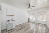 Located at 854 S Oxford Ave in the Koreatown neighborhood of Los Angeles, this one-bedroom, one-bathroom unit offers a clean, minimalist aesthetic defined by an all-white interior and light wood-style vinyl flooring throughout. The main living area is open and airy, featuring a ceiling fan, large windows dressed with slatted blinds that invite natural light, and a compact kitchenette equipped with a two-burner cooktop, sink, overhead cabinetry, and a mini refrigerator — a practical and efficient use of space. A sliding closet door provides built-in storage directly off the main area, complemented by two closets accessible through the unit's corridor. The hallway leads to the generously sized bedroom, which benefits from its own windows and continues the bright, light-filled atmosphere found throughout. Decorative arched door framing adds a subtle architectural character to the interior walls. The bathroom features white tile wainscoting, a pedestal sink with chrome fixtures, a wall-mounted mirror, and a clean, straightforward layout. The unit's consistent white walls and pale flooring create a cohesive, neutral canvas well-suited to a variety of personal styles.