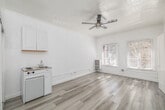 Located at 854 S Oxford Ave in the Koreatown neighborhood of Los Angeles, this one-bedroom, one-bathroom unit offers a clean, minimalist aesthetic defined by an all-white interior and light wood-style vinyl flooring throughout. The main living area is open and airy, featuring a ceiling fan, large windows dressed with slatted blinds that invite natural light, and a compact kitchenette equipped with a two-burner cooktop, sink, overhead cabinetry, and a mini refrigerator — a practical and efficient use of space. A sliding closet door provides built-in storage directly off the main area, complemented by two closets accessible through the unit's corridor. The hallway leads to the generously sized bedroom, which benefits from its own windows and continues the bright, light-filled atmosphere found throughout. Decorative arched door framing adds a subtle architectural character to the interior walls. The bathroom features white tile wainscoting, a pedestal sink with chrome fixtures, a wall-mounted mirror, and a clean, straightforward layout. The unit's consistent white walls and pale flooring create a cohesive, neutral canvas well-suited to a variety of personal styles.