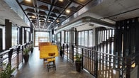 The Music Room at 1601 Broadway in San Diego presents a compelling blend of industrial and contemporary design across a single floor. The space is defined by its striking coffered ceiling, finished in dark metal paneling with recessed lighting that casts a warm, focused glow throughout. Exposed silver HVAC ductwork runs prominently overhead, reinforcing the industrial aesthetic alongside raw concrete walls and polished concrete floors.

Expansive floor-to-ceiling glazing lines the perimeter, flooding the interior with natural light and providing panoramic views of the surrounding San Diego streetscape. Dark-framed metal railings with perforated mesh panels add a refined industrial edge, separating circulation areas while maintaining visual openness throughout the space.

A vintage drafting table and stool serve as a notable furnishing element, nodding to a creative, studio-like atmosphere. Tall potted plants are positioned throughout, offering organic contrast to the hard industrial materials. A separate enclosed room with glass-panel doors and a pass-through observation window provides an acoustically distinct area, likely suited for recording or monitoring purposes. The overall ambiance is creative, urban, and purposefully designed for music or arts-oriented use.