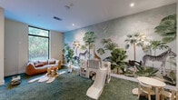 Located in North Miami Beach, FL, this imaginative children's playroom is designed to inspire creativity and active play. The space is defined by a breathtaking floor-to-ceiling safari mural that spans an entire wall, depicting a lush tropical landscape complete with giraffes, zebras, an ostrich, exotic birds, and towering palm trees — bringing the wonders of the natural world indoors in vivid detail.

The room features plush green carpet throughout, creating a soft and safe surface for play. A variety of play equipment is thoughtfully arranged across the space, including a white toddler slide-and-climb structure, wooden balance boards, rocking animal toys, and small child-sized seating. A wall-mounted flat-screen television is positioned at an appropriate height for young viewers, while built-in storage shelving helps keep toys and supplies organized.

Whimsical wicker animal wall décor — including a giraffe and elephant — adds a charming, nature-inspired aesthetic that complements the mural. Recessed lighting and warm wall sconces provide a well-lit environment, while large floor-to-ceiling windows along one wall flood the room with natural light and offer leafy outdoor views. The clean white walls and modern finishes round out a space that is both functional and delightfully enchanting for children.