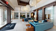 Omnia at Town Center presents a thoughtfully designed amenity floor that combines sophistication with everyday functionality. The grand lobby welcomes residents with soaring double-height ceilings, large-format white tile flooring, and a dramatic cluster of oversized woven globe pendant lights suspended above plush teal velvet seating arrangements. Rich dark wood accents and a bold geometric patterned feature wall add visual depth and elegance throughout the space.

Adjacent to the main lobby, a well-appointed lounge area provides direct access to the outdoor pool deck through expansive glass doors, seamlessly blending indoor and outdoor living. A casual seating area with vibrant orange dining chairs offers a relaxed space for socializing.

The private dining room is a standout feature, designed for refined gatherings with a live-edge wood dining table, upholstered chairs, dark stone accent walls, integrated cabinetry, and ambient cove lighting overhead. Nearby, a fully equipped kitchen includes stainless steel double wall ovens and a touchscreen automated coffee station, catering to residents' culinary and entertaining needs.

Smaller private workspaces are available, finished with marble-style tile walls and warm wall sconces, ideal for focused work or meetings. The fitness center rounds out the amenities with a full suite of modern strength and cardio equipment, floor-to-ceiling mirrors, and large windows providing natural light and neighborhood views.