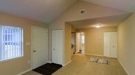This single-floor residence offers a welcoming and functional layout with a blend of warm, neutral tones and thoughtful design elements throughout. The entryway is finished with dark hardwood-style flooring, providing a clean transition point into the home, and is flanked by a white paneled front door and a bifold closet door offering convenient storage from the moment you walk in. The vaulted ceiling in the entryway area adds a sense of openness and grandeur, drawing the eye upward and enhancing the overall sense of space. Moving into the main living area, plush carpeting creates a comfortable, cozy atmosphere underfoot. The open layout connects the living space seamlessly to the rest of the home, with a clear sightline leading through to the kitchen and an adjacent room beyond. The kitchen area, visible in the background, benefits from a window that introduces natural light, brightening the interior. Throughout the space, neutral beige walls provide a versatile backdrop, complemented by white trim and baseboards that give a clean, finished look. Additional closet space is accessible in the living area, adding practicality to the home's design. The residence presents a balanced combination of comfort, storage, and open living space.