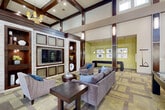 Located in Woodbridge, Virginia, this single-floor community clubhouse offers a generously sized, open-concept layout that seamlessly blends a fully equipped kitchen with a spacious living and lounge area. Soaring ceilings with dramatic dark wood coffered beams define the architectural character of the space, lending both grandeur and warmth throughout.

The kitchen is appointed with sleek dark cabinetry, striking white marble-veined countertops, and a vibrant green mosaic tile backsplash that adds a pop of color. A large center island with bar seating provides an ideal gathering spot, while stainless steel appliances and under-cabinet lighting complete the modern, functional design. Large windows allow natural light to pour in, brightening the space.

The living area is equally impressive, featuring a patterned carpet floor in earthy gold and taupe tones that anchors multiple seating arrangements, including plush sofas, armchairs, and café-style bistro tables. A built-in entertainment wall with stacked stone accents, open shelving, and a large flat-screen TV serves as the focal point of the lounge. A pool table anchors the central gathering zone, reinforcing the space's social and recreational purpose. Floor-to-ceiling windows with elegant drapery frame outdoor views and flood the interior with light, creating a welcoming and vibrant community atmosphere.