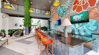Located at the Promenade in Doral, FL, this generously sized single-floor amenity lounge offers a bold and energetic atmosphere designed for social living. The space is anchored by dramatic floor-to-ceiling columns wrapped in cascading live greenery, which draw the eye upward to the double-height ceiling and gold-accented pendant lighting suspended throughout.

A striking hand-painted tropical mural dominates the feature wall, showcasing vibrant florals and monstera leaves in coral, teal, and slate tones — setting a lively, resort-inspired aesthetic. Beneath it, a sleek bar counter with geometric tile detailing is lined with orange barstools and equipped with mounted flat-screen displays, making it ideal for both social gatherings and casual work sessions.

The lounge area is furnished with a mix of modern sofas, woven accent chairs, and bold curved red booth seating arranged around intimate tables. A central billiards table serves as a focal point for recreation and entertainment. The light-toned tile flooring with contrasting inlaid patterns adds visual texture throughout.

Large open-air passages connect seamlessly to an outdoor courtyard featuring palm trees, resort-style seating, and a pool area, allowing natural light and tropical breezes to flow freely into the interior. The overall design blends lush biophilic elements with vibrant contemporary style.