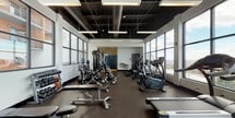This single-floor fitness center is designed with a modern industrial aesthetic, characterized by an exposed black ceiling that highlights structural elements and ductwork overhead. Suspended rectangular light fixtures provide consistent, even illumination throughout the space, complementing the abundant natural light that streams in through expansive floor-to-ceiling windows lining multiple walls. These large-pane windows offer open views of the surrounding urban and natural landscape, creating an energizing atmosphere for workouts.

The flooring consists of durable rubber matting, practical for both heavy equipment and high-traffic use. The layout is thoughtfully organized, with a dedicated cardio zone featuring a row of treadmills, elliptical machines, and stationary bikes positioned near the windows. A separate strength-training area includes a multi-station cable machine, free weight benches, barbells, and a well-stocked dumbbell rack. The open floor plan allows for easy circulation between zones.

Toward the rear of the space, a bank of lockers in teal tones provides convenient storage, alongside a water fountain station. White walls keep the interior bright and clean, balancing the bold industrial ceiling and creating a focused, professional fitness environment overall.