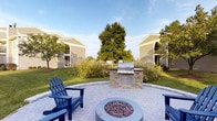 This outdoor communal space is thoughtfully designed for relaxation and entertaining. At its center sits a circular paver patio finished in a classic brick-tone pattern, anchoring the area with a round fire pit filled with decorative lava rock. Blue Adirondack chairs flank the fire pit, providing comfortable seating with a welcoming, casual feel. A built-in outdoor kitchen station constructed with stacked stone detailing houses a stainless steel multi-burner gas grill, offering a dedicated space for outdoor cooking and gatherings. The patio is framed by neatly maintained green lawn and ornamental plantings, including natural grasses and mature trees that provide shade and a sense of privacy. The surrounding residential buildings feature neutral siding and arched architectural detailing, contributing to a cohesive and polished aesthetic. The open sky above and abundant greenery create a bright, airy atmosphere ideal for outdoor leisure. The space balances functional amenities with attractive landscaping, making it a practical and inviting environment for social gatherings throughout warmer seasons.