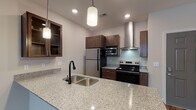 Located at 9921 Horizon Overlook Dr in Raleigh, NC, this single-floor residence offers a well-designed layout with thoughtful finishes throughout. The kitchen is a standout feature, boasting granite countertops with an undermount sink, rich dark-walnut cabinetry — including glass-front upper cabinets — and a full suite of stainless steel appliances including a refrigerator, electric range, built-in microwave, and a sleek stainless range hood. Pendant lighting and hardwood-style flooring complete the kitchen's polished look. The open living area transitions seamlessly from the kitchen, featuring neutral carpeting and access to an outdoor balcony through a set of French doors topped with a transom window, inviting natural light into the space. The bedroom is comfortably sized, accented by a ceiling fan and windows with blinds that allow for natural light and outdoor views. A spacious walk-in closet with wire shelving provides generous storage. The bathroom is finished with large-format tile flooring, a granite-topped vanity with dark cabinetry, and a full soaking tub with shower. The overall aesthetic is clean and contemporary, with neutral tones creating a calm and functional living environment.