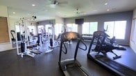This single-floor fitness center is thoughtfully designed to support a comprehensive workout experience. The space is outfitted with a strong selection of cardio equipment, including multiple treadmills positioned near large windows that flood the room with natural light and provide open, airy views to the exterior. A wall-mounted flat-screen television adds convenience for those looking to stay entertained during cardio sessions.

The strength training area is well-appointed with a variety of cable and resistance machines, offering options for a full-body workout. A dedicated free weights section lines the perimeter, featuring a neatly organized dumbbell rack with a wide range of weights to accommodate all fitness levels.

The flooring consists of durable, dark rubber matting suited for high-traffic gym use, while ceiling fans overhead ensure proper air circulation throughout the space. Large mirrors adorn one wall, amplifying the sense of space and allowing users to monitor their form during exercise.

Recessed lighting complements the abundant natural light from the windows, keeping the environment bright and motivating. The overall ambiance is clean, functional, and well-maintained, making this fitness center a practical and inviting space for daily exercise routines.