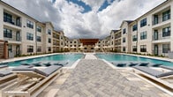 Located at 23615 Kingsland Blvd in Houston (Katy), TX, this outdoor amenity space showcases a beautifully designed resort-style pool area that serves as a central gathering hub for the community. The expansive swimming pool is the focal point, featuring crystal-clear blue water, built-in tanning ledges with in-water lounge chairs, and a sun shelf that invites relaxation. The pool is framed by a generously sized stone-paved sundeck finished in a neutral, multi-toned paver pattern that complements the upscale aesthetic of the surroundings.

A wood-beamed pergola structure anchors one end of the space, providing shaded seating and an outdoor lounge area with a warm, inviting atmosphere. Umbrella-shaded seating clusters are distributed throughout the deck, offering multiple areas for lounging and socializing. Lush accent plantings are strategically placed along the perimeter, softening the hardscape and adding greenery to the open-air environment.

The pool area is enclosed by modern multi-story buildings featuring white board-and-batten and brick facades, large black-framed windows, and private balconies that overlook the pool below. The architectural design reflects a contemporary farmhouse style that blends clean lines with traditional materials, creating a cohesive and visually appealing outdoor retreat.