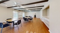 Located at 555 E Bonita Ave in San Dimas, CA, this single-floor community space offers a well-appointed layout designed for group gatherings and shared use. The main gathering area is the heart of the space, characterized by striking dark exposed ceiling beams that add architectural character and warmth. Recessed lighting, flush-mount fixtures, and dual ceiling fans ensure the room remains bright and comfortable. Warm hardwood-style flooring runs throughout, complementing the neutral, beige-toned walls. Multiple tables with seating are arranged throughout the open room, making it well-suited for meetings, events, or communal activities. Large vertical-blind windows along the walls allow natural light to fill the space and offer views of the surrounding area. A built-in shelving unit provides additional display or storage functionality along one wall. The fully equipped kitchen features rich cherry wood cabinetry, granite-style countertops, a gas range, refrigerator, and a double sink beneath a window — ideal for catering or community meal preparation. Two bathrooms round out the amenities, supporting the space's function as a versatile, full-service gathering facility. Overall, the space presents a clean, inviting, and functional environment suitable for a wide range of community or organizational activities.