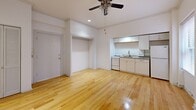 Located at 1103-1117 SW Alder St in Portland, Oregon, this studio unit offers an efficient and thoughtfully arranged single-floor layout. The combined kitchen and living area forms the heart of the space, finished with warm, light-toned hardwood floors that extend throughout and reflect natural light to create a bright, inviting atmosphere. A ceiling fan with integrated lighting anchors the living area, complementing recessed lighting overhead. Two windows fitted with blinds allow natural light to filter in while offering privacy. The kitchen is outfitted with white cabinetry, granite-style countertops, a double sink, and white appliances including a range and refrigerator. A wall-mounted air conditioning unit and a wall heater provide climate control year-round. A dedicated closet offers convenient storage, while a short hallway connects the main living area to the full bathroom. The bathroom is cleanly finished with tile surround, a soaking tub and shower combination, a pedestal sink, and light wood-tone flooring. The unit's crisp white walls and clean lines throughout give the space a tidy, move-in-ready character well-suited to urban living in downtown Portland.