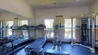This athletic center is a dedicated fitness facility designed to support a well-rounded cardiovascular workout routine. The space is outfitted with multiple treadmills, an elliptical cross-trainer, and additional exercise equipment, providing users with a variety of cardio and strength training options. Large windows line the walls, flooding the room with natural light and offering views of the surrounding landscaped greenery, creating an open and energizing atmosphere. Full-length mirrors are mounted along one wall, enhancing the sense of space and allowing users to monitor their form during workouts. The flooring features a durable, cushioned surface in a deep blue tone, well-suited for high-traffic fitness use. The walls are finished in a neutral tone, contributing to a clean and focused environment. An athletic center rules and policies notice is posted on the wall, indicating that this facility is part of a managed property or community. The ceiling features recessed lighting and a ventilation system, ensuring the space remains comfortable during use. Overall, the athletic center is a practical and functional fitness space that offers a pleasant workout environment with ample natural light and essential cardio equipment.