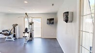 Located at 201 Idlewild Rd in Bel Air, Maryland, this fitness center offers a generous, open single-floor layout designed to accommodate a comprehensive range of workout activities. The space is finished with durable rubber-style flooring throughout, providing a safe and functional surface ideal for both strength training and cardio exercises.

The exercise room is expansive and well-organized, housing a robust selection of professional-grade equipment. Strength training is well represented with Hoist-brand cable machines, a Smith machine with an adjustable bench, a leg press, and a dumbbell rack stocked with a full range of free weights. Cardio offerings include treadmills, ellipticals, and a stationary bike, all positioned to take advantage of the natural light streaming through large windows that overlook the outdoor surroundings.

A bold geometric accent wall in teal and charcoal adds a dynamic visual focal point to the space, complementing the clean white walls and recessed ceiling lighting that keep the environment bright and energizing. Wall-mounted televisions are positioned for convenient viewing during workouts. A dedicated closet provides additional storage for accessories and equipment. The overall ambiance is clean, modern, and purposefully designed to support a full fitness routine.