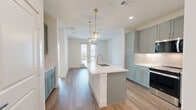 Located in McKinney, TX, this well-appointed one-bedroom, one-bathroom residence offers a thoughtfully designed single-floor layout with modern finishes throughout. The open-concept kitchen and living area serves as the heart of the home, featuring warm wood-look flooring, crisp white walls, and abundant natural light. The kitchen stands out with sage-green shaker cabinetry, quartz countertops, a large undermount sink, gold-accented hardware and faucets, stainless steel appliances including a French door refrigerator, electric range, built-in microwave, and dishwasher, as well as a stylish subway tile backsplash and elegant pendant lighting over the island. The living area opens to a private balcony through glass French doors, offering outdoor views of the surrounding community. The bedroom is carpeted for comfort, equipped with a ceiling fan, and features a large window that brings in natural light. It connects directly to the walk-in closet, which includes built-in shelving and a hanging rod for organized storage. The bathroom is finished with matching sage cabinetry, quartz counters, gold fixtures, a framed mirror, and a glass-enclosed walk-in shower, all carried out in a cohesive, contemporary aesthetic.