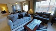 This single-floor residence offers a comfortable and well-appointed living environment that blends style with functionality. The living area serves as the heart of the home, featuring plush blue-gray sofas accented with decorative throw pillows, a rich dark wood coffee table with a glass inset top, and a patterned area rug that anchors the seating arrangement. Large windows dressed with vertical blinds and teal curtain panels flood the space with abundant natural light, creating a bright and airy atmosphere throughout the day. An elegant arched architectural detail adds a decorative focal point to the wall, lending a refined character to the interior. The living space transitions seamlessly into the dining area, which is illuminated by a warm pendant light hanging above a dark wood dining table and chairs, perfect for everyday meals or entertaining guests. The combination of hardwood-style and carpeted flooring further defines the distinct yet connected zones within the open layout. Accent lamps and decorative accessories are thoughtfully placed throughout, contributing to a warm and inviting ambiance. Overall, this residence presents a cohesive, well-lit interior that balances practical living with an appealing aesthetic sensibility.