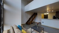 This two-floor loft-style space is defined by its dramatic double-height ceilings that create an immediate sense of openness and volume upon entry. The main living area on the ground floor features polished concrete flooring and clean white walls, lending the space a contemporary, industrial aesthetic. A comfortable seating area anchors the living room, complemented by a textured area rug and a sleek dark coffee table that add warmth and contrast to the minimalist surroundings. A standout architectural feature is the open-tread staircase with rich wood steps and black metal railings, which serves as both a functional and design centerpiece, connecting the ground floor to the upper level. The upper floor overlooks the living area through an open balustrade, reinforcing the loft's sense of verticality and flow. Adjacent to the living space, a kitchen area is visible with light-toned cabinetry and a countertop pass-through, offering a functional layout that integrates smoothly with the open-plan design. Recessed and overhead lighting throughout ensures the space is well-illuminated. The overall ambiance is modern and urban, balancing industrial elements with warm finishes to create a versatile and visually dynamic living environment.