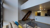 This two-floor loft-style space is defined by its dramatic double-height ceilings that create an immediate sense of openness and volume upon entry. The main living area on the ground floor features polished concrete flooring and clean white walls, lending the space a contemporary, industrial aesthetic. A comfortable seating area anchors the living room, complemented by a textured area rug and a sleek dark coffee table that add warmth and contrast to the minimalist surroundings. A standout architectural feature is the open-tread staircase with rich wood steps and black metal railings, which serves as both a functional and design centerpiece, connecting the ground floor to the upper level. The upper floor overlooks the living area through an open balustrade, reinforcing the loft's sense of verticality and flow. Adjacent to the living space, a kitchen area is visible with light-toned cabinetry and a countertop pass-through, offering a functional layout that integrates smoothly with the open-plan design. Recessed and overhead lighting throughout ensures the space is well-illuminated. The overall ambiance is modern and urban, balancing industrial elements with warm finishes to create a versatile and visually dynamic living environment.