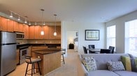 Located at 1982 Route 70 East in Cherry Hill, New Jersey, this single-floor residence offers a well-designed open-concept layout that seamlessly connects the kitchen, dining, and living areas. The kitchen is a standout feature, showcasing warm wood cabinetry, dark granite countertops, and a central island with bar seating, illuminated by elegant pendant lighting overhead. A full suite of appliances and a deep sink complete the functional yet stylish cooking space. The open layout flows naturally into the dining area, which comfortably accommodates a table and chairs, and further into the living room, where plush carpeting and neutral-toned walls create a warm, inviting atmosphere. A framed piece of wall art adds a decorative accent to the living space. The bedroom, visible through a connecting hallway, offers a private retreat with its own furnishings and soft lighting. Throughout the space, the consistent use of neutral tones, recessed lighting, and clean architectural lines contribute to a cohesive and modern aesthetic. The overall design prioritizes both comfort and functionality, making this Cherry Hill residence a well-appointed and welcoming home.