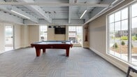This single-floor game room is designed as a dedicated recreational space, offering a well-rounded selection of entertainment options within an open, industrial-style interior. The exposed ceiling structure, finished in light grey, reveals structural beams, ductwork, and linear LED lighting, lending the space a modern yet relaxed atmosphere. Pendant lights with blue shades hang directly over the pool tables, providing focused task lighting while adding a stylish accent to the room.

Two full-sized pool tables serve as the centerpiece of the space, positioned beneath the pendant fixtures and surrounded by ample room for comfortable gameplay. A wall-mounted flat-screen television is installed between large, multi-pane windows, which flood the interior with natural light and offer views of the surrounding exterior. An Imperial shuffleboard table runs along one wall, while a foosball table occupies an adjacent area, rounding out the gaming amenities.

Patterned carpet tiles in a neutral grey-blue tone cover the floors throughout, adding texture and warmth underfoot. A pub-height round table with metal stools provides casual seating for spectators or socializing. A wall-mounted mini-split HVAC unit ensures climate control. The overall ambiance is lively and functional, well-suited for communal recreation.