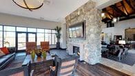 This single-floor space makes an immediate impression with its dramatic floor-to-ceiling river rock fireplace, which serves as the undeniable focal point of the main living area. The fireplace surround, composed of rounded, natural stones, extends upward to the ceiling and is crowned by a large wall-mounted flat-screen television, seamlessly blending rustic craftsmanship with modern convenience. Rich, dark hardwood flooring runs throughout, anchoring the space with warmth and a sense of solidity. The open layout is thoughtfully furnished with rustic wooden tables and chairs alongside upholstered accent seating in earthy, striped tones, creating a comfortable and cohesive aesthetic. Generous windows along one wall invite abundant natural light, offering views to the outdoors and giving the interior a bright, airy quality despite its grounded, lodge-inspired design. Tall potted tropical plants add lush greenery throughout, softening the natural stone and wood elements. Recessed lighting and decorative wall sconces provide balanced illumination, while the high ceiling enhances the sense of spaciousness. The overall ambiance is warm and welcoming, combining natural materials with thoughtful design to create a space that feels both functional and inviting.