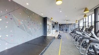 This fitness center offers a comprehensive workout environment designed to accommodate a wide range of exercise activities. The standout feature is a full-height bouldering wall adorned with colorful climbing holds set against a textured gray panel surface, paired with thick protective crash mats on the floor below — an uncommon and impressive amenity that sets this facility apart.

The space extends into a well-equipped cardio and strength training zone, where a row of treadmills and other cardio machines lines one side, while a dedicated strength training area with cable machines and weight equipment occupies the rear. Yellow ceiling pendant lights add a modern, energetic accent throughout, complementing the recessed lighting that ensures the entire space is brightly illuminated.

The interior design incorporates a mix of gray textured wall panels, light carpeted flooring with yellow boundary lines, and large glass partitions that create an open, connected feel. A ceiling fan aids in air circulation over the cardio zone. The overall aesthetic is contemporary and purposeful, balancing both functionality and visual appeal for an engaging fitness experience.
