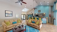 This single-floor community clubhouse is designed with an open-concept layout that seamlessly connects the lounge and kitchen areas into one expansive, welcoming space. The soaring vaulted ceilings create an impressive sense of height and airiness, complemented by a skylight that floods the interior with natural light. Dual ceiling fans provide comfortable circulation throughout the space.

The lounge area is generously appointed with comfortable tan sofas accented by vibrant teal and chartreuse throw pillows, arranged around a dark wood coffee table atop a bold teal area rug. A flat-screen television is mounted on one wall, while framed artwork adds a decorative touch throughout. Large sliding glass doors on one side open to views of an outdoor pool area, enhancing the connection between indoor and outdoor living.

The kitchen area is defined by a striking teal accent wall and features a granite-topped bar counter with chartreuse bar seating. It is equipped with stainless steel appliances, including a full-size refrigerator and microwave, along with ample cabinetry. Pendant lighting over the counter adds a stylish finishing touch.

Tile flooring runs throughout the space, offering both durability and a clean aesthetic. A glass-panel entry door with an arched transom window allows additional natural light to filter in, contributing to the overall bright and inviting atmosphere of this well-designed communal facility.