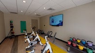 This Fitness On Demand studio is a thoughtfully designed, single-floor exercise space built to support a wide variety of workout routines. The room features durable wood-look flooring accented by a bold yellow mat zone that defines the primary cycling area, where a row of TRUE brand spin bikes are neatly arranged for group or individual cycling sessions. A large wall-mounted flat-screen display at the front of the room streams on-demand fitness classes, providing guided instruction for users at any fitness level. The space is finished with a drop tile ceiling outfitted with recessed lighting, creating a bright and energizing environment. Along one wall, a ceiling-mounted TRX suspension training rig offers dual suspension straps for functional strength and bodyweight training. Additional equipment includes stackable aerobic step platforms, kettlebells of varying weights, medicine balls on a dedicated rack, and resistance bands, ensuring versatility for cardio, strength, and flexibility training. A large glass door at the rear of the studio provides an outdoor view and natural light, with a glimpse of what appears to be a pool area beyond. The neutral, warm-toned walls and clean layout contribute to a focused and motivating atmosphere well-suited for both individual workouts and small group fitness sessions.