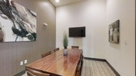 This single-floor meeting space is designed with functionality and modern aesthetics in mind. At the center of the room sits a substantial solid wood conference table with a rich, warm finish, surrounded by matching wooden chairs that comfortably seat a small group — making it well-suited for intimate meetings, presentations, or collaborative work sessions.

A wall-mounted flat-screen display is prominently positioned on the crisp white wall, providing a clear focal point for presentations and video conferencing. The elevated ceiling height lends the room a sense of openness and airiness, preventing the space from feeling confined despite its focused footprint.

The design palette balances warm and neutral tones throughout. One wall features a textured wood-panel accent, adding depth and visual interest, while an abstract art piece introduces a subtle creative touch. A decorative tall grass arrangement in a ceramic vase serves as a natural centerpiece on the table, softening the overall atmosphere.

The flooring consists of a patterned area rug layered over hardwood-style flooring, contributing texture and warmth underfoot. Recessed lighting provides even, ambient illumination throughout the space. Overall, the room strikes a balance between professional utility and refined design, creating an environment conducive to productive engagement.