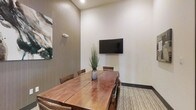 This single-floor meeting space is designed with functionality and modern aesthetics in mind. At the center of the room sits a substantial solid wood conference table with a rich, warm finish, surrounded by matching wooden chairs that comfortably seat a small group — making it well-suited for intimate meetings, presentations, or collaborative work sessions.

A wall-mounted flat-screen display is prominently positioned on the crisp white wall, providing a clear focal point for presentations and video conferencing. The elevated ceiling height lends the room a sense of openness and airiness, preventing the space from feeling confined despite its focused footprint.

The design palette balances warm and neutral tones throughout. One wall features a textured wood-panel accent, adding depth and visual interest, while an abstract art piece introduces a subtle creative touch. A decorative tall grass arrangement in a ceramic vase serves as a natural centerpiece on the table, softening the overall atmosphere.

The flooring consists of a patterned area rug layered over hardwood-style flooring, contributing texture and warmth underfoot. Recessed lighting provides even, ambient illumination throughout the space. Overall, the room strikes a balance between professional utility and refined design, creating an environment conducive to productive engagement.