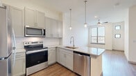 This single-floor unit offers a thoughtfully designed layout that maximizes its compact footprint across a living room, kitchen, bathroom, and closet. The open-concept kitchen serves as a central feature, outfitted with gray shaker-style cabinetry, white subway tile backsplash, and granite countertops. Stainless steel appliances — including a refrigerator, electric range, over-the-range microwave, and dishwasher — provide a complete, modern cooking setup. Two globe pendant lights hang above the peninsula, adding stylish task lighting, while a stainless steel sink with a pull-down faucet sits at the kitchen's corner junction.

The living area is bright and open, benefiting from natural light through a large window with horizontal blinds and a ceiling fan with integrated lighting. Wood-style plank flooring extends throughout, lending warmth and visual continuity to the space. An entry area features an electrical panel and smart thermostat along a clean white wall.

The bathroom is well-appointed with large-format tile flooring, a granite-topped vanity with gray cabinetry, a full tub-shower combination surrounded by white subway tile, and recessed lighting. A dedicated closet provides additional storage. The overall aesthetic is clean, contemporary, and functional.