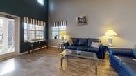 Located at 200 Lakeview Park Rd in Colonial Heights, Virginia, this single-story community clubhouse offers a well-rounded set of amenities designed for both relaxation and activity. The main lounge area makes an immediate impression with its soaring vaulted ceilings and warm hardwood-style flooring. Generous windows flood the space with natural light, while a bold teal accent wall adds a striking visual contrast to the neutral gray surroundings. Comfortable seating and glass-topped tables create an inviting gathering atmosphere. Adjacent to the lounge, a dining area features a glass-top table with seating for four, framed by large windows and a set of French doors that open to the outdoors, offering pleasant views of the surrounding tree-lined grounds. A dedicated workstation area with a private office alcove provides a quiet space for administrative or personal use. The fully equipped kitchen includes stainless steel appliances — refrigerator, range, and over-the-range microwave — complemented by crisp white cabinetry and granite-look countertops. The fitness center is well-appointed with rubber flooring and multiple cardio machines, including treadmills, ellipticals, and a stationary bike, all situated in a bright room with natural light from multiple windows and the same teal accent wall tying the space together cohesively.