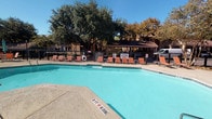 This amenity space features two distinct areas designed to support an active lifestyle. The outdoor pool area is generously sized, with clear turquoise water and a surrounding deck lined with terracotta-colored lounge chairs for sunbathing and relaxation. A covered pavilion structure provides shaded seating, while mature trees frame the perimeter, offering natural privacy and a pleasant outdoor atmosphere. The pool area is enclosed by a wrought iron fence and includes safety signage and equipment.

Adjacent to the pool, the fitness center is a well-lit, single-level space finished with patterned carpet tile flooring and a bold color-block accent wall in orange and blue. Large windows along one wall offer direct views of the pool deck, allowing natural light to fill the space. The gym is equipped with multiple treadmills, elliptical machines, upright and recumbent stationary bikes, a rowing machine, a multi-functional cable and weight machine, and a full dumbbell rack displayed in front of a wall-length mirror. A ceiling fan provides ventilation, and recessed lighting supplements the natural light. The layout accommodates both cardio and strength training efficiently within the space.