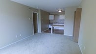 This single-floor, one-bedroom, one-bathroom unit at Bayberry offers a practical and functional layout designed for comfortable everyday living. The living area features neutral-toned carpeting that extends throughout, providing a soft, cohesive feel to the space. Warm beige walls create a calm, understated ambiance throughout the unit.

The kitchen is positioned adjacent to the living area and is defined by a raised counter that serves as a natural dividing element between the two spaces. White upper cabinetry provides ample storage, while the countertops offer functional workspace. A stove and sink are integrated into the kitchen layout, and the flooring transitions to a hard surface, distinguishing the kitchen from the carpeted living area.

A doorway visible from the main living space leads to a secondary area, likely a hallway connecting the bedroom and bathroom, reinforcing the unit's efficient single-floor layout. Overhead lighting fixtures provide illumination throughout. The overall design is clean and straightforward, offering a move-in-ready space suited for individuals or couples seeking a low-maintenance, well-organized home.