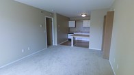 This single-floor, one-bedroom, one-bathroom unit at Bayberry offers a practical and functional layout designed for comfortable everyday living. The living area features neutral-toned carpeting that extends throughout, providing a soft, cohesive feel to the space. Warm beige walls create a calm, understated ambiance throughout the unit.

The kitchen is positioned adjacent to the living area and is defined by a raised counter that serves as a natural dividing element between the two spaces. White upper cabinetry provides ample storage, while the countertops offer functional workspace. A stove and sink are integrated into the kitchen layout, and the flooring transitions to a hard surface, distinguishing the kitchen from the carpeted living area.

A doorway visible from the main living space leads to a secondary area, likely a hallway connecting the bedroom and bathroom, reinforcing the unit's efficient single-floor layout. Overhead lighting fixtures provide illumination throughout. The overall design is clean and straightforward, offering a move-in-ready space suited for individuals or couples seeking a low-maintenance, well-organized home.