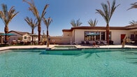 This two-story community amenity space is designed with a distinct Southwestern aesthetic, characterized by stucco exteriors, clay tile rooflines, and warm earth-toned finishes that complement the desert landscape. The centerpiece of the outdoor area is a generously sized, crystal-clear swimming pool with a connected spa, both finished in vivid aquamarine tile that catches the abundant desert sunlight. The expansive pool deck is paved in light-colored stone pavers, providing ample space for lounging and entertaining.

Tall, mature date palms frame the pool area, offering a resort-like atmosphere while providing natural shade throughout the day. The deck is furnished with multiple chaise lounges and shaded seating areas beneath teal market umbrellas and covered pergolas accented with stone columns. Large windows on the adjacent clubhouse structure overlook the pool deck, creating a seamless indoor-outdoor connection.

Access to the pool area is secured through a gated metal fence system with stone-accented walkways and desert landscaping featuring ornamental grasses and decorative gravel. The overall ambiance is bright, open, and recreational, well-suited for a sun-drenched climate and designed to maximize outdoor enjoyment year-round.