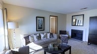 This single-floor space centers around a well-appointed living room that combines comfort and functionality. The room features neutral gray walls and a textured ceiling that lend a calm, understated atmosphere throughout. Plush, light-toned carpeting covers the floor, adding warmth underfoot and contributing to the room's inviting feel.

The living area is anchored by a generously sized sofa adorned with an assortment of decorative pillows in coordinating gray and brown tones. A sleek, dark coffee table sits in front of the seating area, styled with decorative books and accents. A fireplace with a clean, dark surround occupies one wall, serving as a natural focal point for the room.

Wall décor includes a framed cityscape artwork and a wall-mounted mirror, adding visual interest and depth. Natural light filters into the space, brightening the neutral palette. A doorway visible from the living area connects to an adjacent room, suggesting a practical, flowing layout. Overall, the space presents a tidy, comfortable environment with a cohesive neutral color scheme throughout.