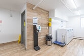 Located at 57 Union St E in Waterloo, Ontario, this shared laundry facility is a compact, efficiently organized space spread across a single floor. The interior is finished with clean white walls, light wood-look flooring, and bright fluorescent tube lighting that keeps the area well-lit throughout.

The space is divided into two interconnected areas. The main laundry room houses multiple front-loading washers and top-loading dryers arranged along the walls, accompanied by a window that brings in natural light. White tile accents and ample machine spacing make for a practical, easy-to-navigate layout.

The adjoining common area features a utility sink set into a white countertop with grey cabinetry below, complemented by a warm wood-paneled accent wall. A self-service payment kiosk is conveniently positioned near the entrance, alongside posted instructions and informational signage. A community bulletin board and a small seating area with an upholstered bench provide a comfortable spot to wait. A washroom is accessible directly off this area via a clearly labeled door.

The overall ambiance is clean, modern, and functional, reflecting a well-maintained shared amenity space designed to serve the needs of its building residents efficiently.