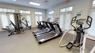 This fitness center is a thoughtfully designed, single-floor space dedicated to health and wellness. The room is bright and welcoming, with an array of large windows framed by classic white shutters and topped with transom windows that allow abundant natural light to fill the space, creating an open and energizing atmosphere.

The facility is equipped with a comprehensive selection of exercise machines to support a full workout routine. Multiple treadmills with integrated digital displays are arranged as the centerpiece of the room, offering a modern cardio experience. Alongside these, additional cardio equipment including rowing machines and an elliptical trainer provide diverse workout options for users of varying fitness levels. A multi-use strength training cable machine rounds out the equipment selection, supporting a range of resistance and strength exercises.

The flooring is a durable, neutral-toned surface well-suited for a high-traffic fitness environment. The drop ceiling features recessed lighting panels that complement the natural light from the windows. An illuminated exit sign and ventilation are also visible, reflecting attention to safety and comfort. The overall ambiance is clean, functional, and inviting — an ideal space for residents or guests seeking a convenient and well-maintained fitness facility.