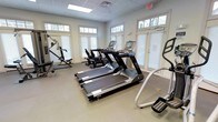 This fitness center is a thoughtfully designed, single-floor space dedicated to health and wellness. The room is bright and welcoming, with an array of large windows framed by classic white shutters and topped with transom windows that allow abundant natural light to fill the space, creating an open and energizing atmosphere.

The facility is equipped with a comprehensive selection of exercise machines to support a full workout routine. Multiple treadmills with integrated digital displays are arranged as the centerpiece of the room, offering a modern cardio experience. Alongside these, additional cardio equipment including rowing machines and an elliptical trainer provide diverse workout options for users of varying fitness levels. A multi-use strength training cable machine rounds out the equipment selection, supporting a range of resistance and strength exercises.

The flooring is a durable, neutral-toned surface well-suited for a high-traffic fitness environment. The drop ceiling features recessed lighting panels that complement the natural light from the windows. An illuminated exit sign and ventilation are also visible, reflecting attention to safety and comfort. The overall ambiance is clean, functional, and inviting — an ideal space for residents or guests seeking a convenient and well-maintained fitness facility.