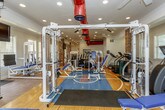 This fitness facility is a thoughtfully designed, single-floor space that blends architectural character with comprehensive workout capabilities. Exposed brick columns serve as defining structural elements throughout, contrasting with crisp white walls and creating a visually dynamic environment. The flooring transitions between warm terracotta tile and a polished hardwood court surface, the latter prominently featuring a "KU" logo at its center, adding a bold, collegiate aesthetic to the space.

The facility is divided into distinct workout zones. One area houses an extensive lineup of Paramount strength-training machines, including cable systems, leg press stations, and adjustable benches, all finished in a white-and-blue color scheme. A dedicated free weights section features a full dumbbell rack positioned along a mirrored wall, maximizing the sense of depth. The cardio zone includes treadmills, stair climbers, and elliptical machines.

Overhead, bold red ductwork runs prominently across the ceiling alongside recessed lighting, contributing to the gym's industrial-inspired character. Multiple windows allow natural light to fill the space, while a wall-mounted television provides entertainment. A ceiling fan aids in air circulation, completing a well-appointed, functional fitness environment.