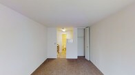 This studio is a single-floor space designed for efficient, straightforward living. The main living area features wall-to-wall carpeting in a neutral tone, providing a comfortable and cohesive foundation throughout the room. The white walls and flat ceiling contribute to a clean, minimalist aesthetic that keeps the space feeling open and easy to personalize.

A built-in closet with bifold doors is conveniently positioned along one wall, offering dedicated storage directly within the living area. Adjacent to the closet, an open doorway provides a seamless transition into the combined kitchen and bathroom zone. The kitchen area is visible from the main room, featuring tile flooring and basic appliances, while the bathroom is situated just beyond, equipped with standard fixtures and overhead lighting.

The layout is straightforward and functional, with each area flowing naturally into the next. The use of light, neutral tones throughout — from the white walls to the light-colored tile in the kitchen and bathroom — helps maintain a sense of continuity and brightness across the space. Overall, this studio offers a practical and well-organized floor plan suited for those seeking a simple, self-contained living environment.
