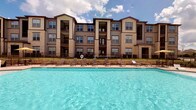 This community amenity space centers around a large, resort-style outdoor pool with crystal-clear blue water, framed by a row of sun loungers and a stone-accented pool deck. The multi-story building backdrop, clad in warm neutral tones with stone masonry detailing and private balconies, adds an upscale architectural presence to the outdoor setting.

Adjacent to the pool, the interior clubhouse offers a welcoming lounge area finished with warm hardwood-style flooring, crown molding, and recessed lighting complemented by ceiling fans. The main gathering room features comfortable leather seating arranged around a wall-mounted television and entertainment unit, alongside multiple round café-style tables and chairs — ideal for socializing or casual dining. The sage-green walls are adorned with contemporary framed artwork, lending a polished, cohesive aesthetic throughout.

A kitchenette serves the space with dark espresso cabinetry, granite countertops, stainless steel appliances, and a striking mosaic tile backsplash illuminated by pendant lighting. Steps lead to an upper-level area, offering additional seating and a dedicated workspace. Together, these amenities create a functional and stylish communal environment designed for relaxation and community engagement.