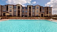 This community amenity space centers around a large, resort-style outdoor pool with crystal-clear blue water, framed by a row of sun loungers and a stone-accented pool deck. The multi-story building backdrop, clad in warm neutral tones with stone masonry detailing and private balconies, adds an upscale architectural presence to the outdoor setting.

Adjacent to the pool, the interior clubhouse offers a welcoming lounge area finished with warm hardwood-style flooring, crown molding, and recessed lighting complemented by ceiling fans. The main gathering room features comfortable leather seating arranged around a wall-mounted television and entertainment unit, alongside multiple round café-style tables and chairs — ideal for socializing or casual dining. The sage-green walls are adorned with contemporary framed artwork, lending a polished, cohesive aesthetic throughout.

A kitchenette serves the space with dark espresso cabinetry, granite countertops, stainless steel appliances, and a striking mosaic tile backsplash illuminated by pendant lighting. Steps lead to an upper-level area, offering additional seating and a dedicated workspace. Together, these amenities create a functional and stylish communal environment designed for relaxation and community engagement.