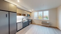 This single-floor unit showcases a thoughtfully designed, contemporary interior with a clean, modern aesthetic throughout. Light wood-tone luxury vinyl plank flooring runs continuously across the space, contributing to a bright and cohesive atmosphere.

The kitchen is a standout feature, equipped with stainless steel appliances including a gas range with built-in display, over-the-range microwave, dishwasher, and refrigerator. Two-tone cabinetry pairs warm natural wood upper cabinets with slate gray lower cabinets, complemented by under-cabinet lighting and a textured white geometric tile backsplash. A farmhouse-style sink with a matte black faucet adds a refined design detail.

A large picture window floods the living area with natural light while offering views of the surrounding neighborhood and tree canopy. A wall-mounted HVAC unit provides climate control. The living space is open and versatile, with neutral white walls and recessed lighting.

A sliding-door closet and dedicated entryway provide practical storage and a clear transition into the unit. The bathroom features a full tub-shower combination with white subway tile surround, matte black fixtures and hardware, a wood-accented vanity with a white countertop, and light wood flooring, completing the modern and cohesive design palette.