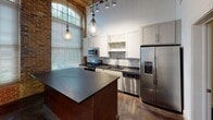 This single-floor loft showcases a compelling blend of industrial heritage and contemporary design. The main living area commands attention with soaring wood-paneled ceilings supported by exposed timber beams, oversized arched windows set within original brick walls, and gleaming polished concrete floors that reflect abundant natural light. A large ceiling fan circulates air throughout the expansive open space, while exposed ductwork adds to the authentic industrial character.

The kitchen is a standout feature, offering white shaker cabinetry, a subway tile backsplash, dark stone countertops, and a full suite of stainless steel appliances including a range, microwave, dishwasher, and refrigerator with ice maker. A substantial island with a dark concrete top provides generous prep and dining space, illuminated by pendant globe lights and track lighting above.

A private room with neutral-toned walls and concrete flooring provides a quieter retreat, framed by an original exposed timber post. The bathroom is finished with a dual-sink vanity featuring dark stone countertops, round mirrors, industrial-style cage light fixtures, and a tiled tub-shower combination. A sliding barn-style door adds a functional and stylish touch to the interior transitions. Overall, the space exudes a warm yet edgy industrial ambiance throughout.