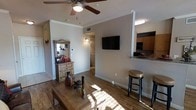 This single-floor space showcases a thoughtfully arranged open layout that connects the living area to the kitchen, creating a cohesive and functional flow throughout. The living area features warm hardwood-style flooring that extends across the space, complemented by crisp white crown molding and neutral-toned walls that lend a clean, inviting atmosphere. A ceiling fan with a light fixture is centrally positioned overhead, providing both airflow and ambient lighting.

A wall-mounted flat-screen television serves as a focal point on the main living room wall, while a rustic wooden dresser with a mirror adds a decorative touch and additional storage in the corner. Decorative accents, including ceramic vases and a star ornament on the coffee table, contribute warmth and character to the room.

The kitchen, partially visible from the living area, features wood-toned cabinetry, granite-style countertops, and a raised breakfast bar with bar stool seating—ideal for casual dining. A hallway leading from the living area provides access to additional rooms beyond. Natural light filters through the space, highlighting the warm tones of the flooring and furnishings throughout.