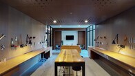 Located at 931 Fletcher Ave in Indianapolis, Indiana, this single-floor space masterfully combines a professional workshop environment with a refined lounge area. The open-plan layout is anchored by polished concrete floors and a distinctive perforated ceiling panel finished in warm wood tones, lending both acoustic character and visual depth to the interior.

The workshop zone features continuous built-in wooden workbenches lining both sides of the room, accented by under-bench ambient lighting. A large live-edge wood communal worktable serves as a central focal point. Pegboard walls display an organized array of tools and adjustable task lighting arms, creating a highly functional and well-equipped workspace.

Transitioning seamlessly into the lounge area, rich warm-toned slatted wood wall paneling frames a wall-mounted flat-screen television, flanked by classic molded shell rocking chairs and a leather sofa. Floor-to-ceiling glazed windows and sliding glass doors flood the space with natural light and offer direct views of the landscaped exterior walkway.

A utility area toward the rear includes a vending machine, a kitchenette with sink, and additional workspace, ensuring convenience for occupants. The overall ambiance is sleek and industrial yet inviting, making this a versatile space suited for creative work, collaboration, and relaxation.